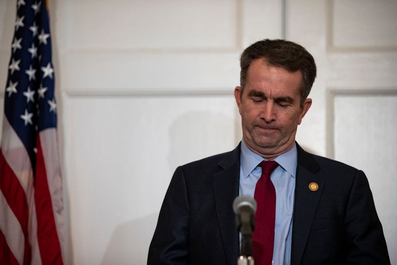 Northam, standing in front of an American flag on a flagpole, looks downward.